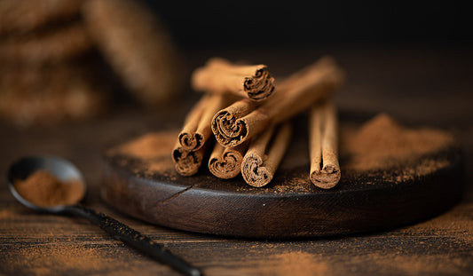 Ceylon cinnamon quills on a wooden plate, with ceylon cinnamon powder dispersed around the cinnamon quills, and cinnamon powder on a spoon next to the wooden plate.